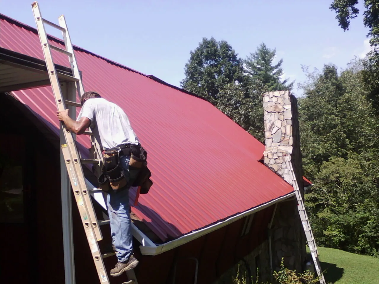 Roofer on a ladder beside a red metal roof and stone chimney — Soffit & Fascia Repair by Roofing Brothers in Altadena